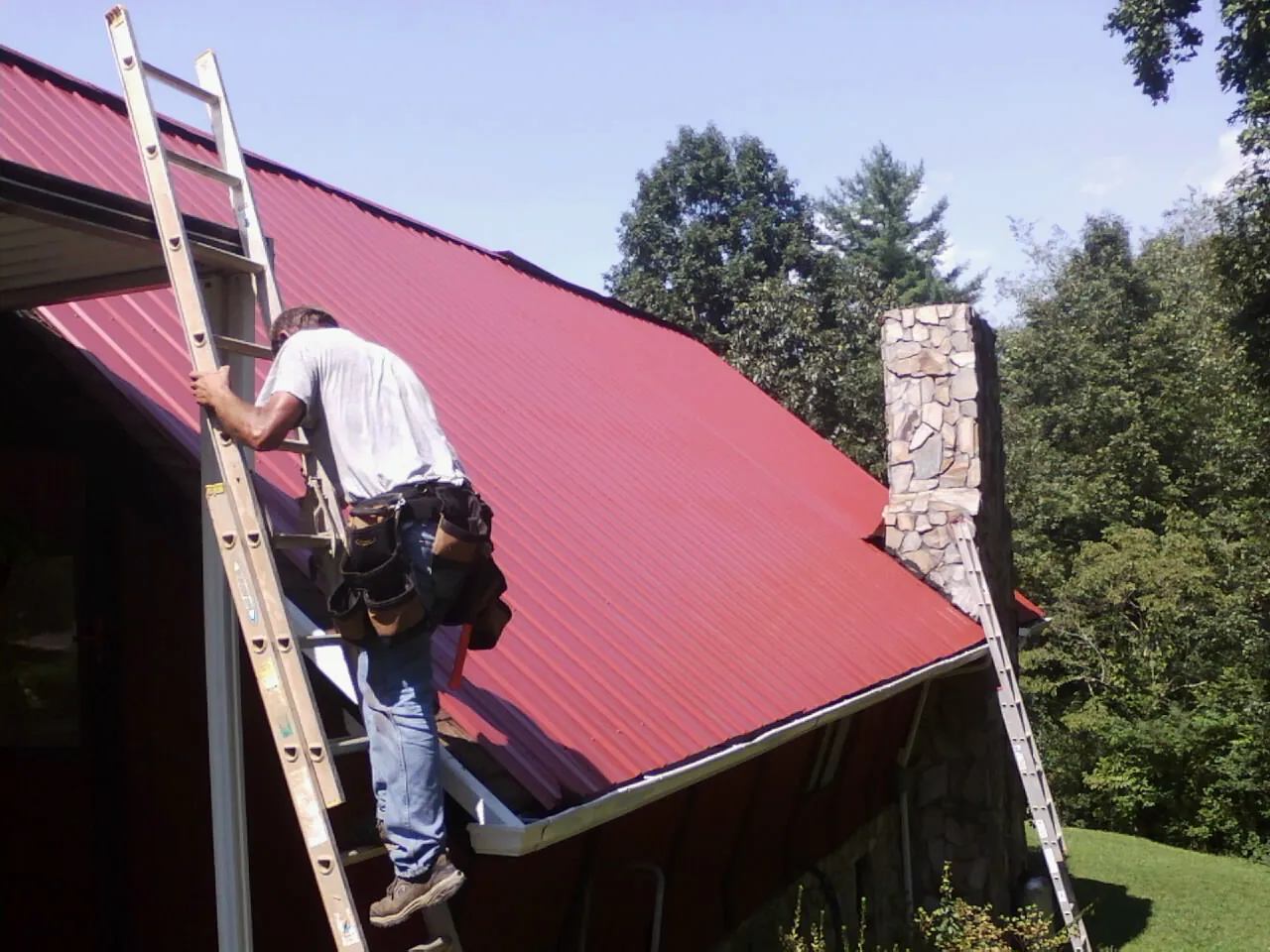 Roofer on a ladder beside a red metal roof and stone chimney — Soffit & Fascia Repair by Roofing Brothers in Rachel